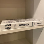 A hardcover sketchbook titled 'Warner Turtle Sketchbook' resting on a white shelf inside a white wooden cabinet or shelf unit.